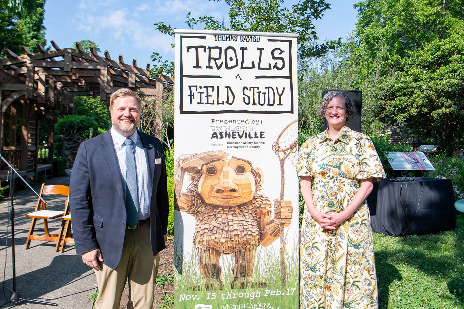 North Carolina Arboretum Executive Director Drake Fowler and Explore Asheville and Buncombe County Tourism Development Authority President and CEO Vic Isley stand next to Trolls: A Field Study banner at Thursday's press conference at the NC Arboretum Heritage Garden