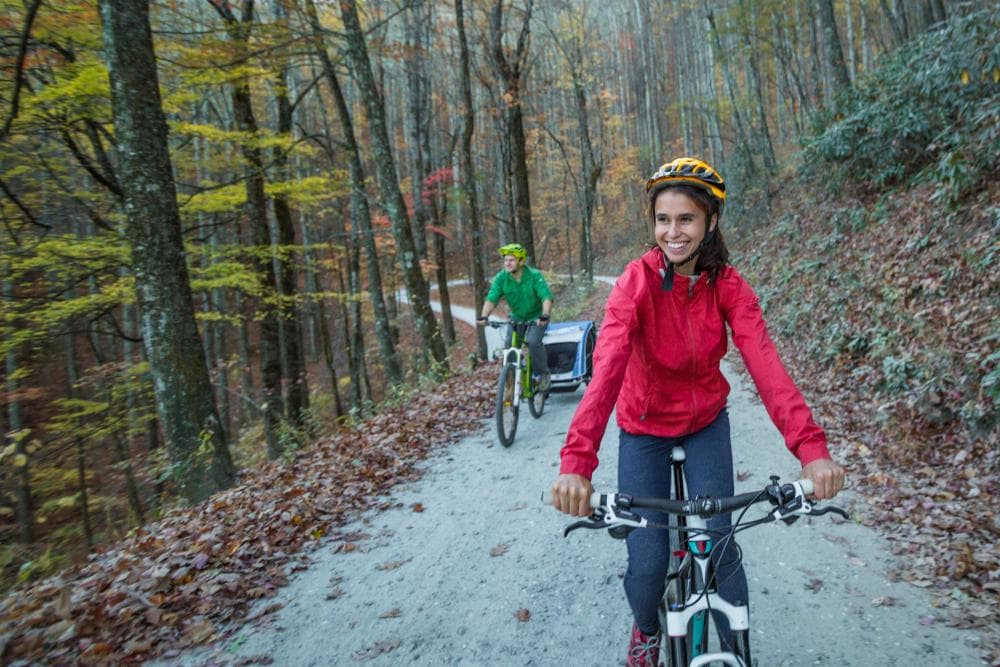 Woman wearing pink jacket riding bike on trail with husband behind during fall Woman wearing pink jacket riding bike on trail with husband behind during fall