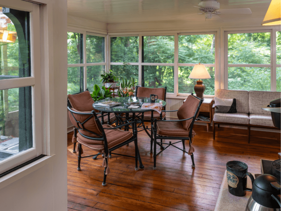 Screened sun porch at Pinecrest Bed & Breakfast in Asheville's Montford Historic District, with cushioned chairs around a glass table and woodland views through wraparound windows Screened sun porch at Pinecrest Bed & Breakfast in Asheville's Montford Historic District, with cushioned chairs around a glass table and woodland views through wraparound windows