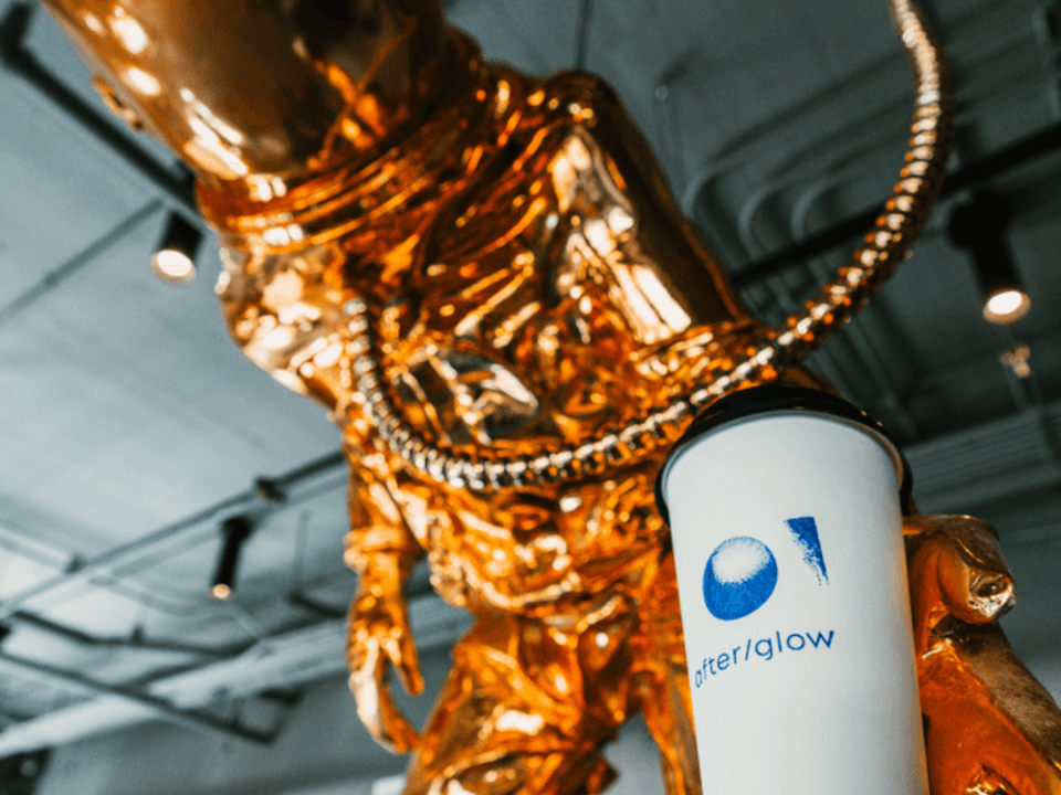 golden astronaut handing the viewer a to-go mug with after/glow stamped on it in blue golden astronaut handing the viewer a to-go mug with after/glow stamped on it in blue