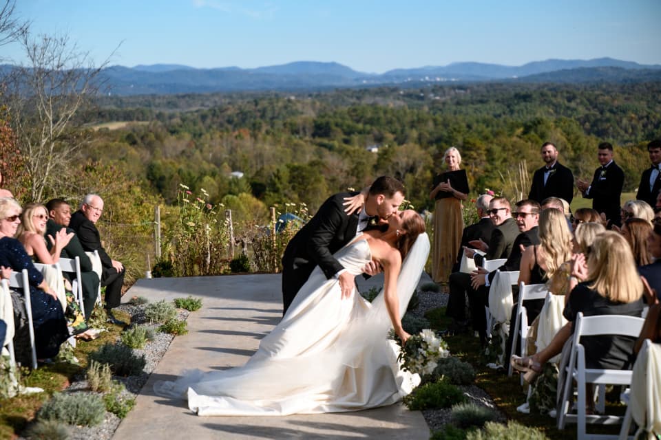 A bride and groom share a dip kiss at an outdoor wedding ceremony at Lonigleaf Vineyard, with guests seated on either side and greenery and the Blue Ridge Mountains in the background. A bride and groom share a dip kiss at an outdoor wedding ceremony at Lonigleaf Vineyard, with guests seated on either side and greenery and the Blue Ridge Mountains in the background.