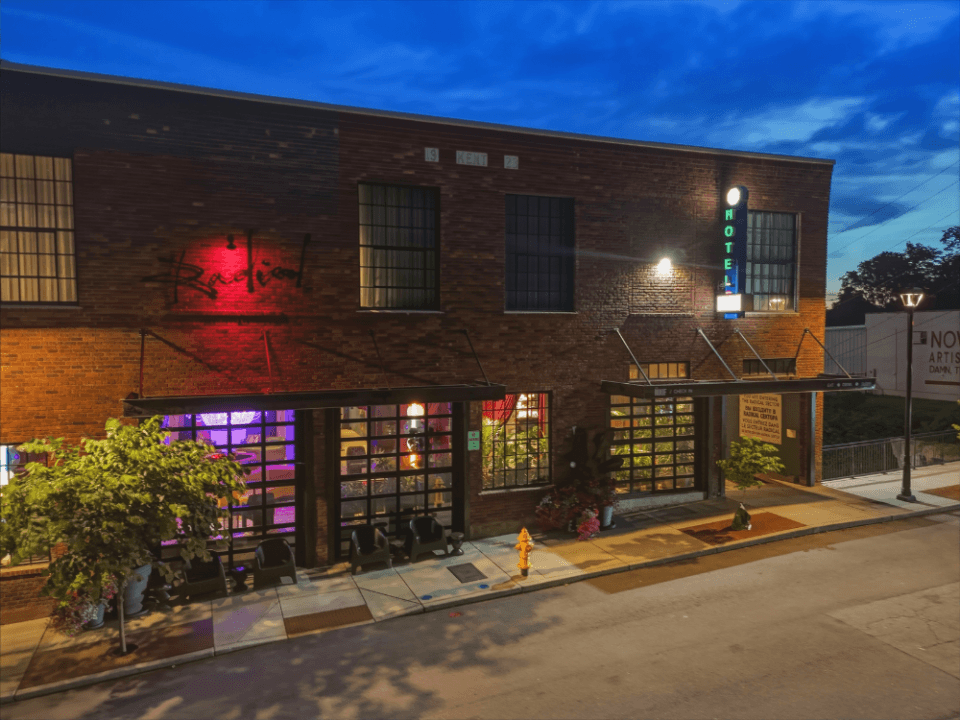 exterior image of The Radical Hotel from the street level at dusk so you can see into the lobby exterior image of The Radical Hotel from the street level at dusk so you can see into the lobby