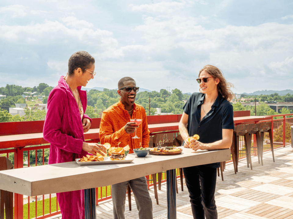 three individuals smiling with sunglasses on eating and drinking on a high top table on a rooftop bar in the sun three individuals smiling with sunglasses on eating and drinking on a high top table on a rooftop bar in the sun