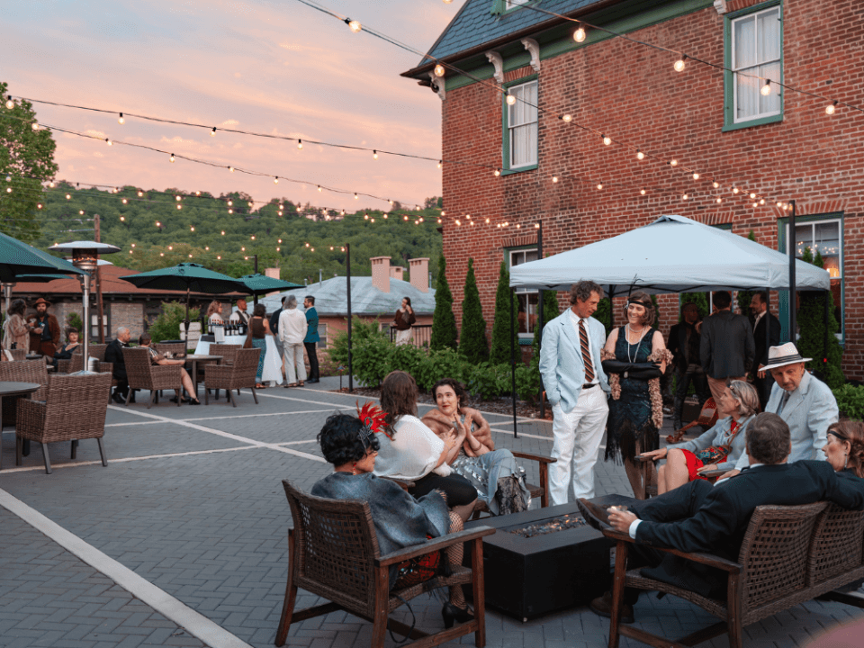 Patio next to brick house with people enjoying sitting outside under string lights and a sunset Patio next to brick house with people enjoying sitting outside under string lights and a sunset