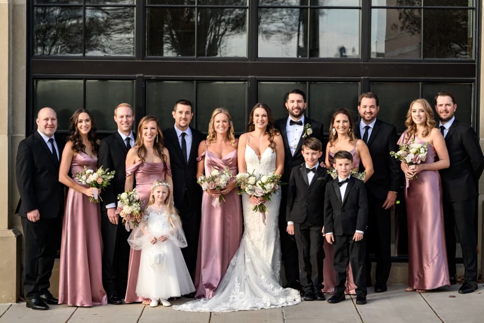 A wedding party poses outside; the bride in white, groom in a tuxedo, bridesmaids in pink dresses, groomsmen in black suits, and two children dressed formally at a Restoration Hotel wedding. A wedding party poses outside; the bride in white, groom in a tuxedo, bridesmaids in pink dresses, groomsmen in black suits, and two children dressed formally at a Restoration Hotel wedding.