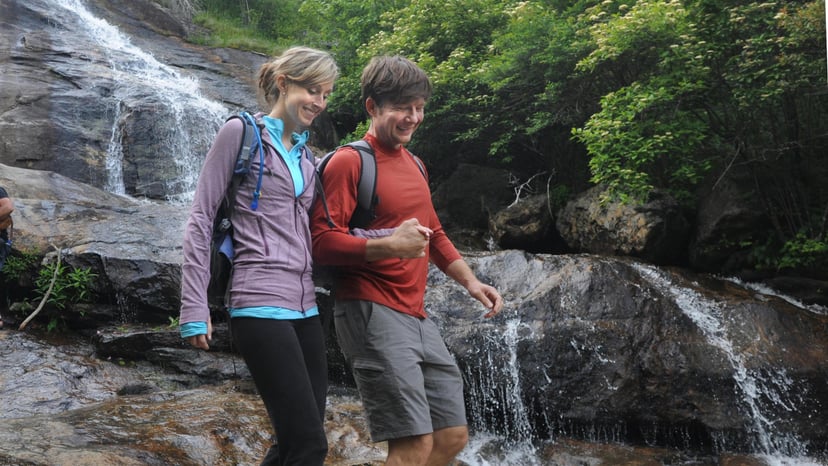 Two adults smile and lock arms as they pass a rock with water cascading down it near Asheville
