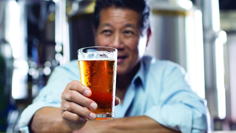 a person in a dress shirt smiles while holding a beer in front of them at a bar
